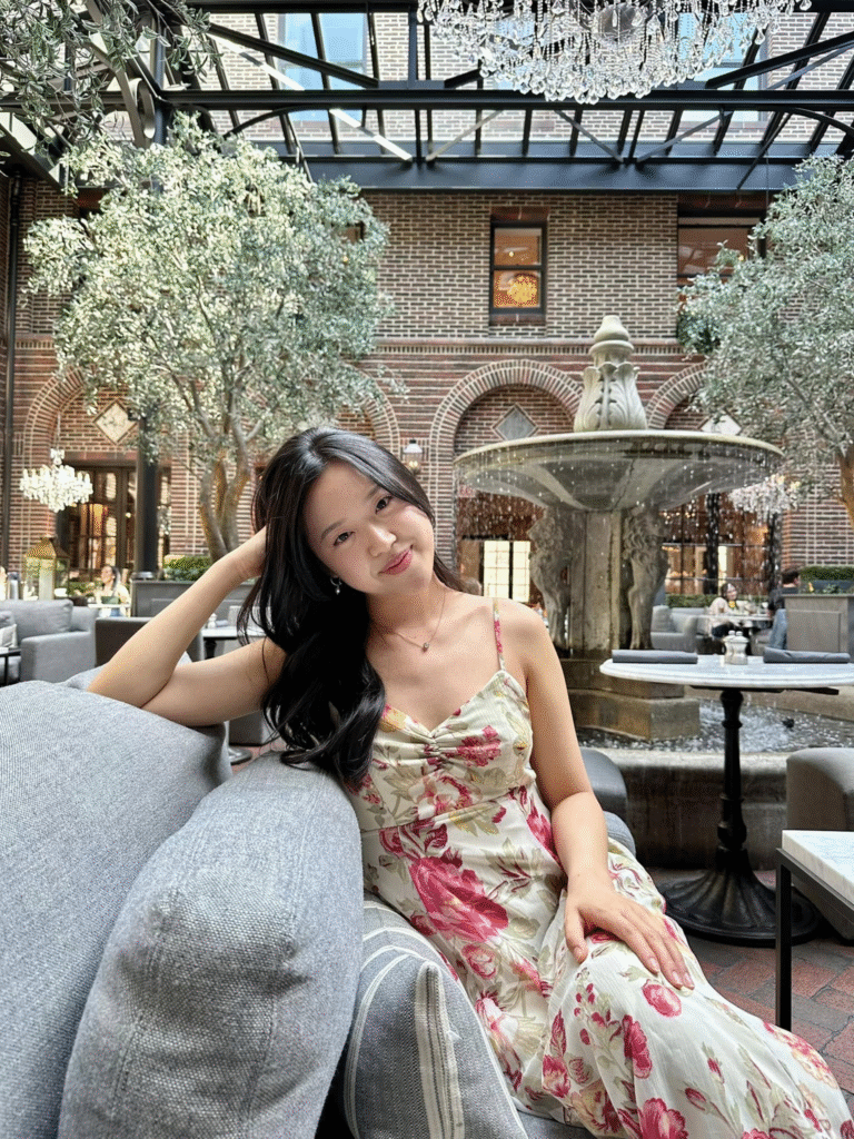 Allison Baek embodies effortless sophistication, posing as the quintessential It Girl in a lush, sun-drenched atrium setting.